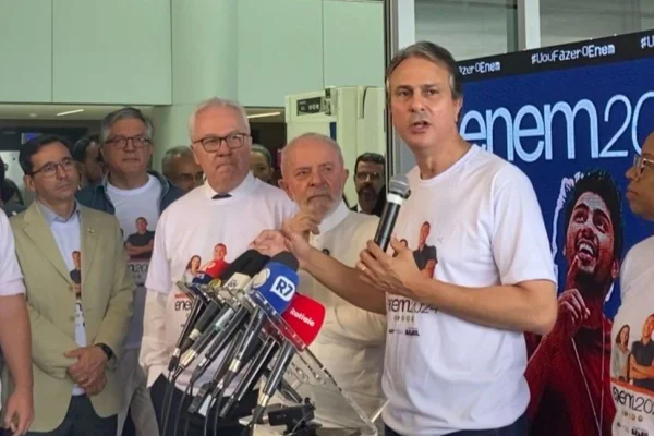 Imagem colorida mostra Camilo Santana, de camiseta branca, discursando sobre o Enem ao lado do presidente Lula - Metrópoles