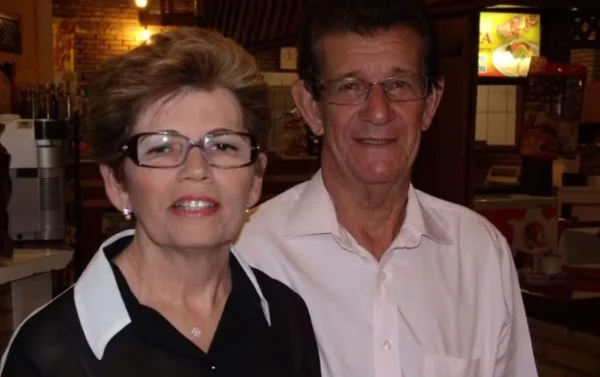 Imagem colorida de casal, mulher tem cabelos curtos castanhos, usa óculos e camisa preta com detalhes brancos. Homem idoso tem cabelo castanhos, usa óculos e veste camisa branca