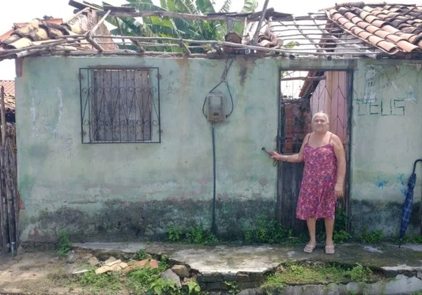 mulher idosa usa um vestido rosa e posa para foto em frente a uma casa humilde e em más condições