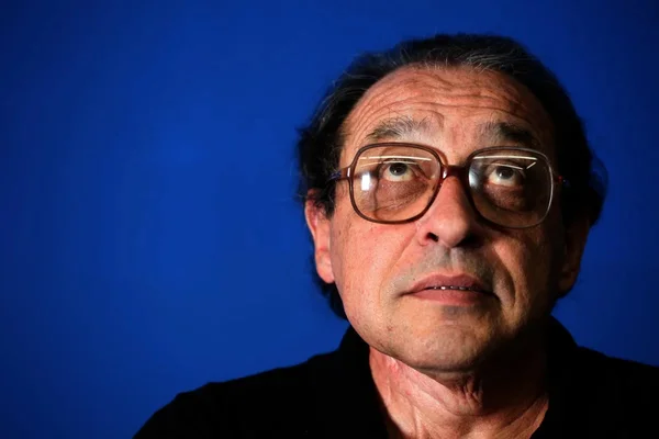 Imagem colorida de Ruy Castro olhando para cima, de óculos, em fundo azul - Metrópoles
