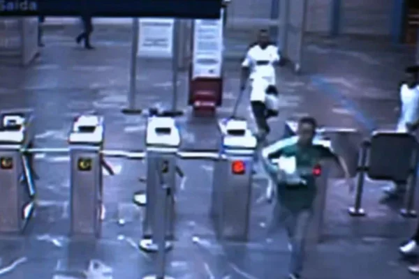 Imagem mostra print de vídeo de câmera de segurança de estação de trem em São Paulo. É possível ver catracas , um homem correndo na frente de outro homem que segura uma barra em uma das mãos - Metrópoles