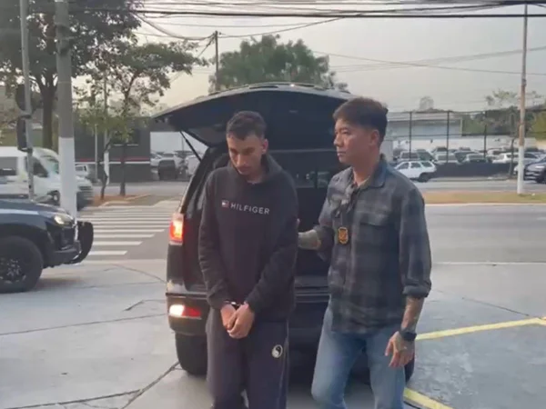 Foto colorida mostra policial conduzindo suspeito algemado da viatura para a delegacia.