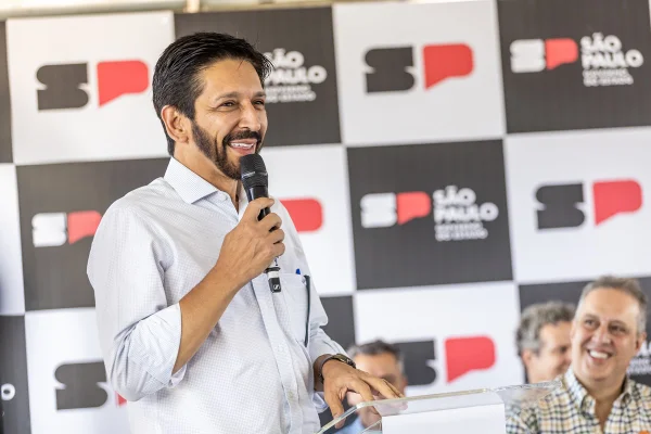 Ricardo Nunes, homem branco, de cabelo e barba pretos, de camisa branca, fala ao microfone na frente de um banner do governo estadual - Metrópoles