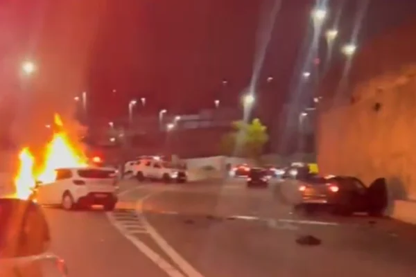 Imagem colorida de carro branco de motorista de app pegando fogo e carro cinza batido no canto direito da avenida. Metrópoles