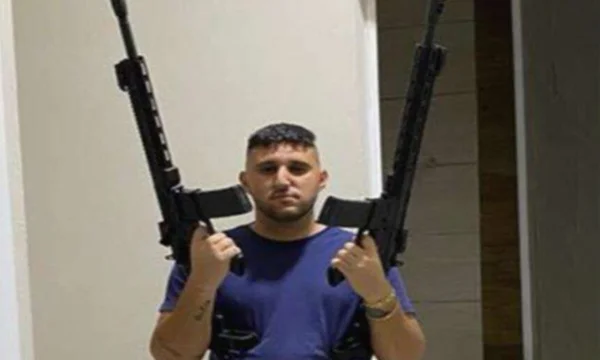 Foto colorida criminoso com o nariz torto usa camiseta azul e segura dois fuzis, um em cada mão - Metrópoles