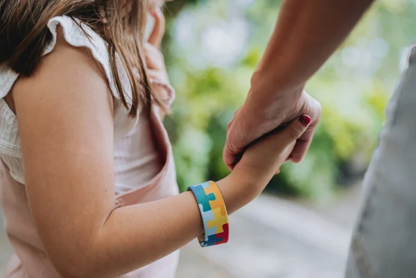 Foto colorida de criança com pulseira símbolo do autismo segurando mão de adulto - Metrópoles