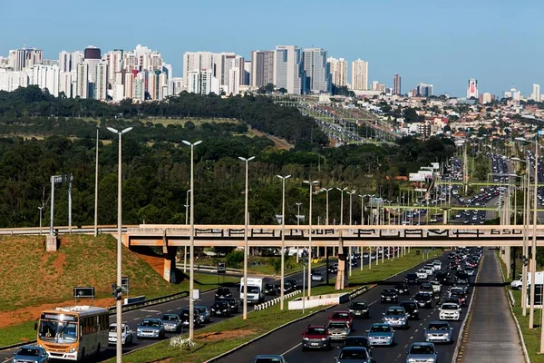 Pista com carros passando e viaduto e prédios ao fundo