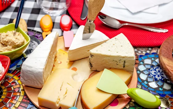 Sobre a mesa com toalha xadrez, vemos sobre uma tábua de vidro alguns queijos, como Gorgonzola, Brie, Provolone, Camembert e Estepe