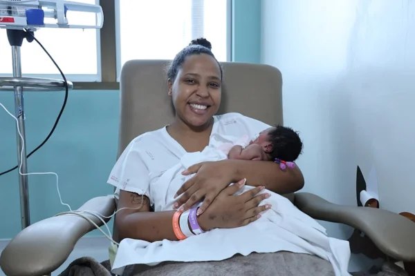 Foto colorida de mulher em ambiente hospitalar segurando uma bebê rencém nascida - Metrópoles.