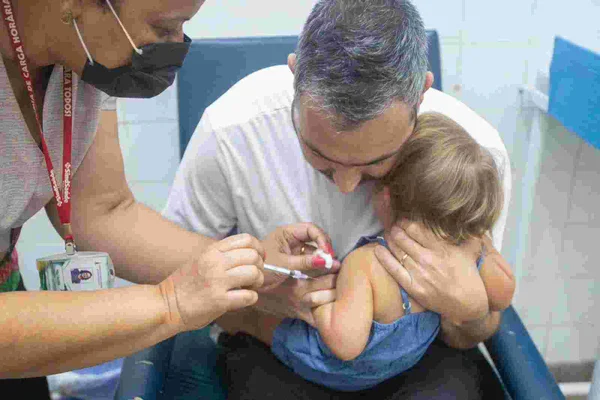 Foto colorida de pai segurando criança para vacinação - Metrópoles
