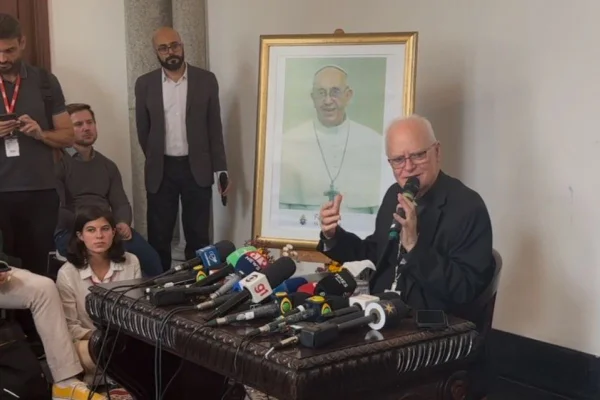 Imagem colorida mostra homem idoso com cabelos brancos e roupa preta de padre. Ele segura um microfone na mão esquerda. Ao fundo há um quadro com foto o papa Francisco - Metrópoles