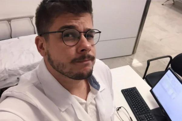 Homem de barba rala e óculos de grau, usando jaleco branco de médico, em consultório - Metrópoles