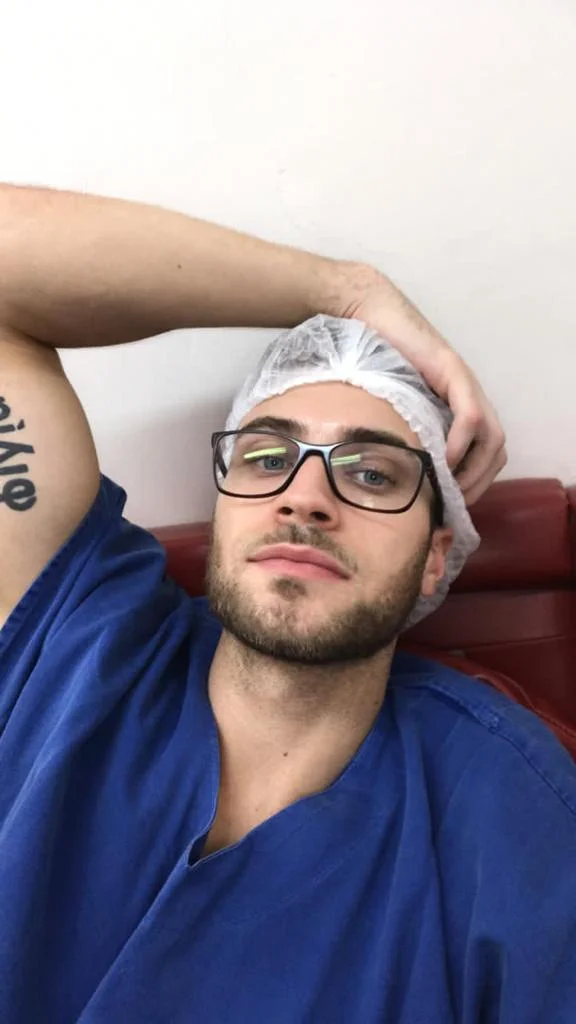 imagem colorida mostra homem loiro de olhos verdes, pele branca, óculos de grau com armação preta usando camiseta azul e touca cirurgica branca - Metrópoles