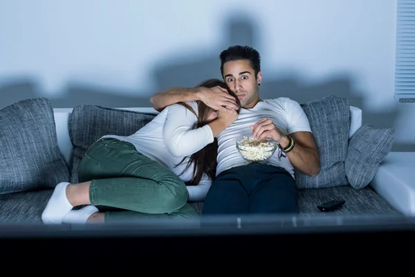 Imagem colorida de casal sentado no sofá assistindo a filme de terror - Metrópoles