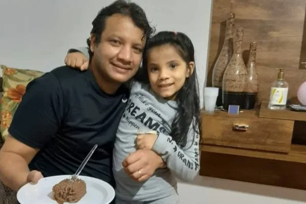 Foto colorida de homem segurando prato, no qual há um pedaço de bolo de chocolate, abraçando uma crianças - Metrópoles