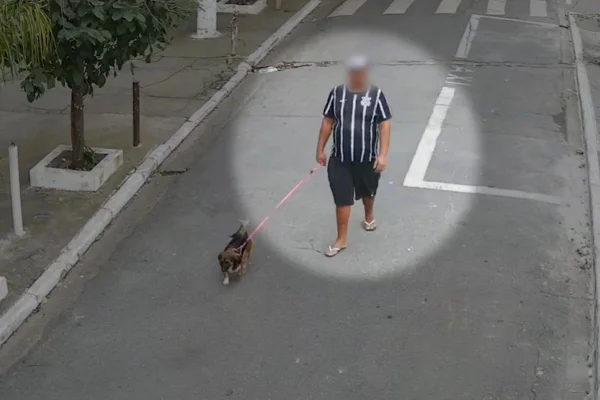 Homem com camisa listrada de time e boné branco caminha pela rua segurando cachorro pela coleira -Metrópoles