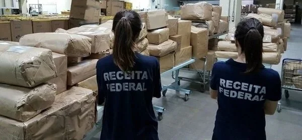 Agentes da Receita Federal durante operação olhando para galpão cheio de caixas sob mesas e no chão. Elas são vistas de costas, uniformizadas e ambas usam rabo de cavalo - Metrópoles