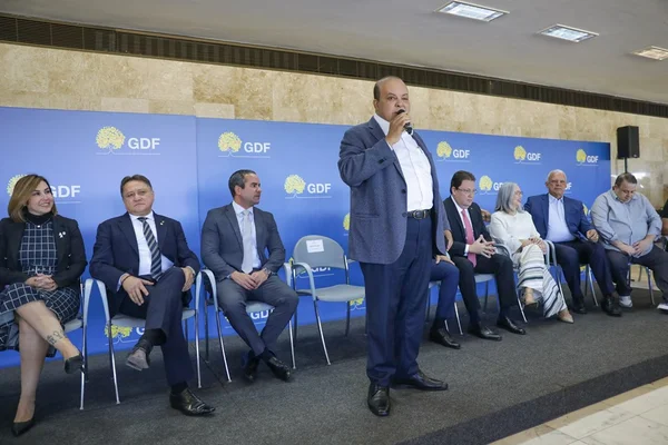 Foto colorida do governador Ibaneis Rocha, em pé, falando ao microfone. Atrás dele, há integrantes do GDF e de parlamentares