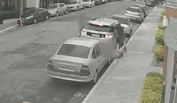 Imagem colorida mostra criminoso empurrando mulher em calçada, próximo a uma fila de carros - Metrópoles