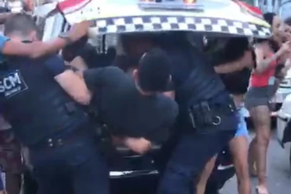 Dois guardas, cada um de um lado, seguram homem pelos braços e tentam coloca-lo dentro de compartimento traseiro de viatura - Metrópoles