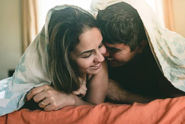 Foto colorida de casal rindo juntos embaixo do cobertor deitados na cama - Metrópoles