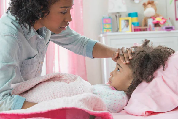 Mãe tocando testa da filha deitada na cama com lençois cor de rosa