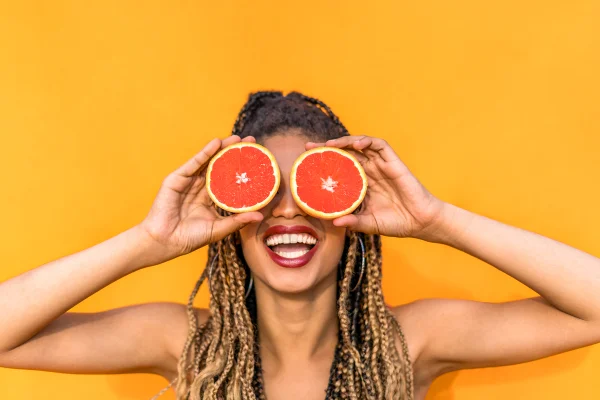 Imagem mostra mulher negra, com cabelo trançado, sorrindo e segurando duas metade de laranja na frente dos olhos - Metrópoles