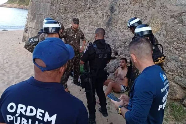 foto colorida de homem sentado preso e sentado em chão de praia com policiais e agentes da SEOP ao redor - metrópoles