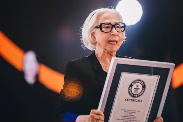 Na imagem colorida, Fernanda Montenegro aparece de roupa preta com óculos de armação grossa. Ela segura placa do Guinness Book, o Livro dos Recordes, e faz discurso - Metrópoles