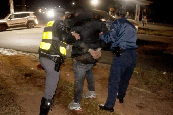 Dois policiais seguram pelos braços um homem algemado. Os três estão de costas - Metrópoles