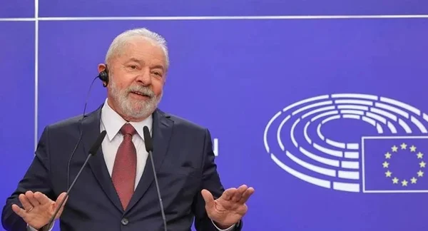 Na imagem colorida, Lula está posicionado do lado esquerdo, sob fundo azul. Ele usa terno escuro, camiseta branca e gravata vermelha. Na frente dele há um microfone e ele está com as mãos levantadas - Metrópoles