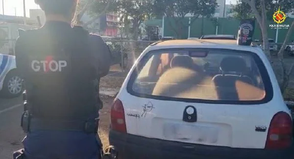 policial militar de costas e diante de um Corsa branco com a placa borrada
