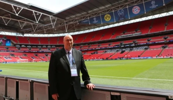 Homem carece usando terno e óculos posando para foto dentro de um estádio de futebol na Europa