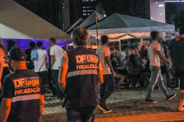 Homens de costas com farda e pessoas em espaço aberto com tendas ao fundo