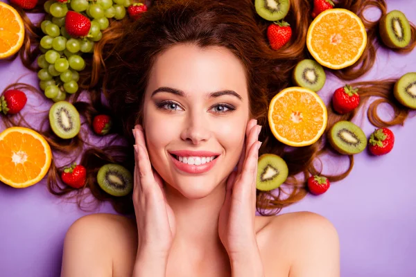 Foto colorida. Imagem mostra mulher branca, ruiva deitada sob um fundo roxo e com várias frutas ao redor - Metrópoes