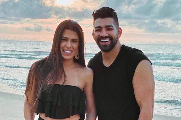 Imagem colorida de Dilsinho e Beatriz Ferraz em praia, ambos usando preto e sorrindo - Metrópoles