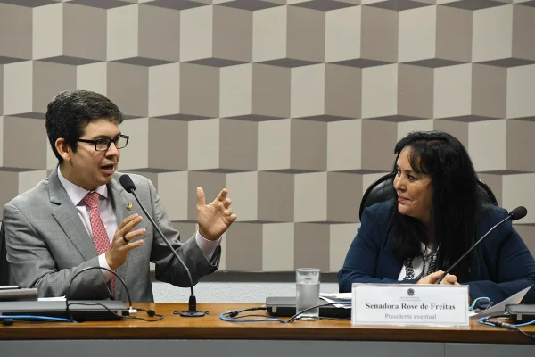 Os senadores Randolfe Rodrigues e Rose de Freitas sentados lado a lado na mesa diretora de uma comissão do Senado. Randolfe fala ao microfone enquanto Rose, sorrindo, o observa - Metrópoles
