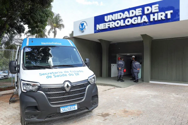 Ambulância branca estacionada em frente à Unidade de Nefrologia HRT. A placa azul com o nome da unidade aparece acima da entrada. Profissionais circulam pelo local