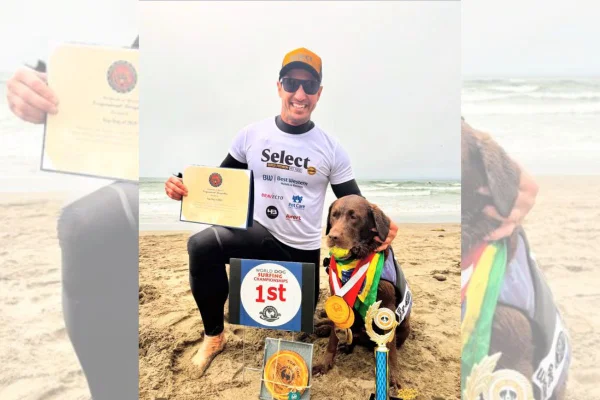 Homem na praia, abraçado com uma labradora com medalhas e troféus