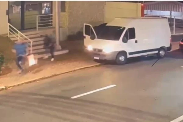 Homem com mala de mão corre em calçado, perseguido por homem de prato, armado com fuzil. Mais atrás, perua branca, com porta do passageiro aberta - Metrópoles