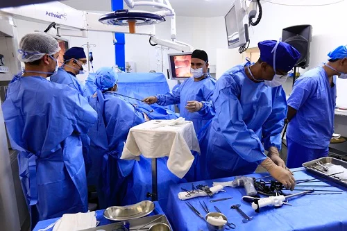 Imagem colorida de uma sala de cirurgia onde profissionais estão usando roupas hospitalares azuis