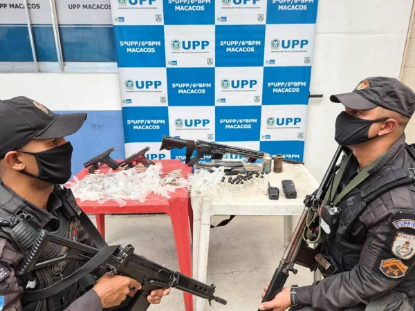 Dois PMs armados aparecem no primeiro plano, no fundo uma mesa com armas e drogas