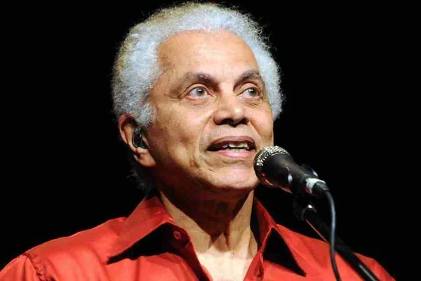 Foto colorida de Paulinho da Viola. Ele está de blusa vermelha e canta em apresentação - Metrópoles
