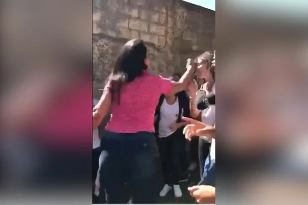 mulher de camiseta rosa com braço estendido atingindo rosto de aluna