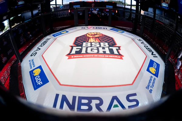 A foto mostra uma luva de boxe branca com logos e, ao fundo, o octógono iluminado em vermelho e azul, com refletores e telões ao redor