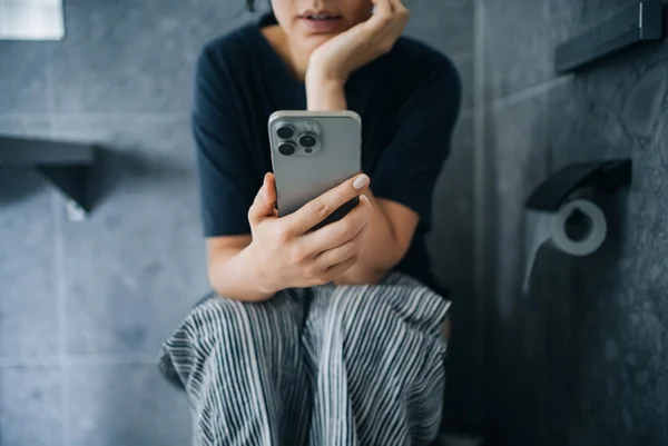 Foto colorida de mulher sentada em vaso sanitário mexendo no celular - Metrópoles