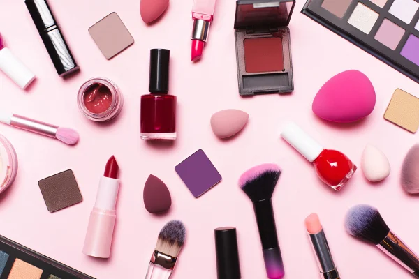 Conjunto de maquiagem com vista superior sobre fundo rosa pastel. Pincéis, batons, pó facial, blushes, esponjas, sombras e esmaltes. Metrópoles