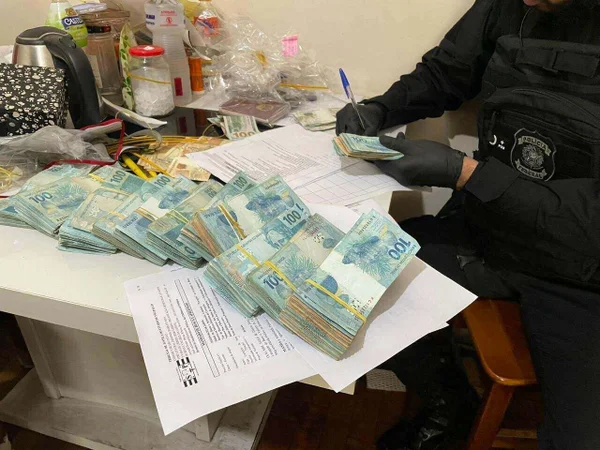 Foto colorida mostra maços de notas de 100 reais sobre mesa enquanto agente da PF faz anotação