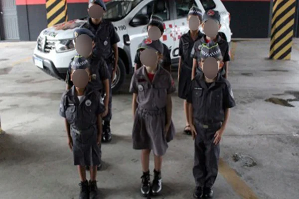 Iamgem colorida mostra oito crianças, com uniformes da PM, enfileiradas na frente de uma viatura - Metrópoles