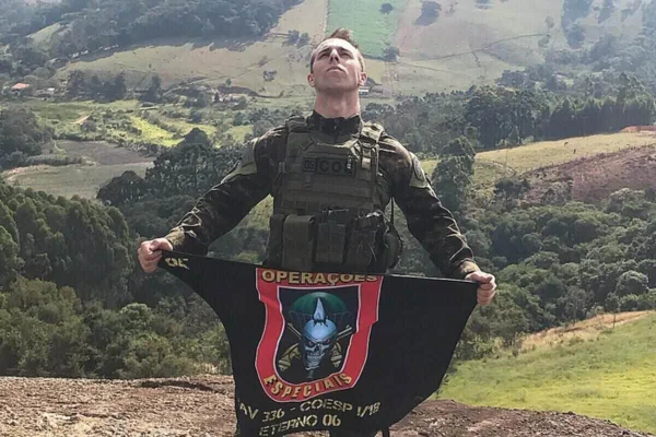 Foto colorida de policial com farda verde escura, em montanha em dia ensolarado, segurando abaixo da cintura uma bandeira do grupo de operações especiais da PM de São Paulo - Metróles